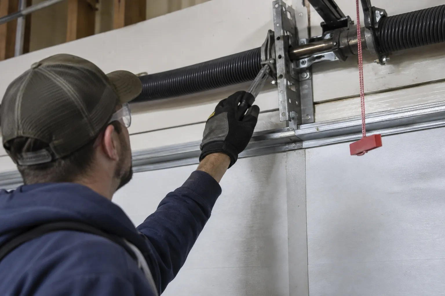 Technician in a cap reaching a gloved hand with a tool toward a coiled spring and bracket above a garage door.